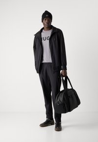 Black athletic jacket, gray shirt with "HUGO" text, matching black pants, and black sneakers. The model holds a black duffel bag.