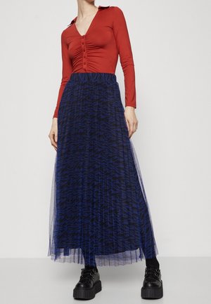 Vrouw draagt een roestrood, geplooid shirt met lange mouwen en knopen, gecombineerd met een blauw-zwart patroon plissé maxirok en zwarte platformschoenen.