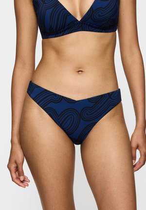 Kvinde iført en blå bikini med sorte abstrakte swirl-mønstre, der viser mellemgulv og hofter mod en ensfarvet baggrund.