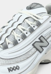 Hvid og grå New Balance 1000 sneaker med mesh-stof, bølge-mønstrede læderoverlayer og hvide snørebånd der er trukket gennem grå løkker.