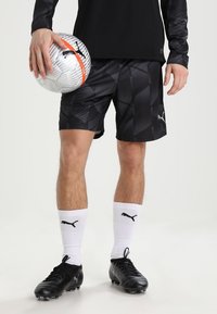 Svarta sportshorts med ett geometriskt mönster, matchat med svarta fotbollsskor och vita strumpor med en svart logotyp. Håller en silver- och orange fotboll.