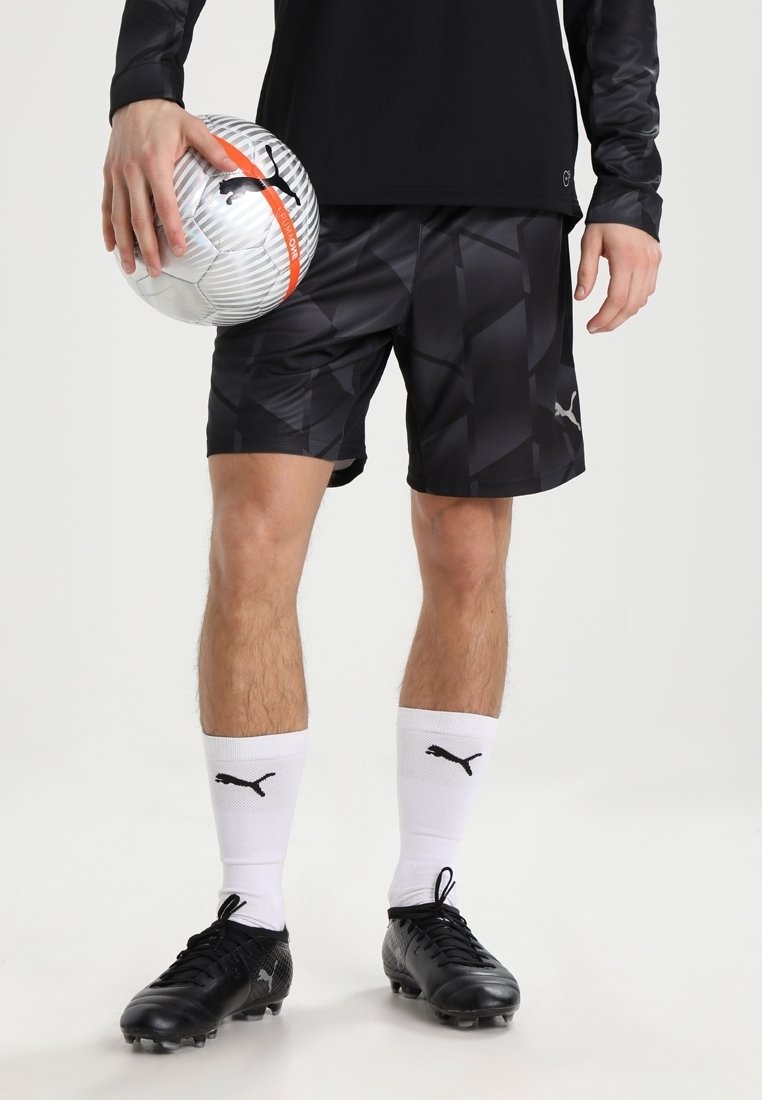 Svarta sportshorts med ett geometriskt mönster, matchat med svarta fotbollsskor och vita strumpor med en svart logotyp. Håller en silver- och orange fotboll.