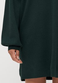 Sötétzöld kötött ruha hosszú, dús ujjaival és enyhén bő szabásával; bordás mandzsettadetalizálással és oldalsó hasítékokkal rendelkezik.