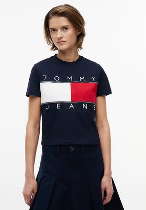 Ung kvinde iført en navyblå Tommy Jeans crop top med et rødt, hvidt og navyblåt logofelt, kombineret med navyblå plisserede bukser.