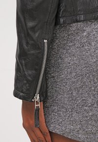 Giacca di pelle nera con manica con zip e hardware argentato, contrastata da un tessuto grigio testurizzato sottostante.