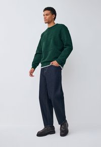 Zielony sweter z tłoczonym wzorem, czarne dżinsy i ciemne obuwie. Sweter ma prążkowane mankiety i okrągły dekolt.