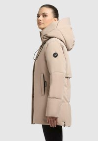 Giacca a vento beige con un grande cappuccio, tasche laterali e dettagli del logo neri. La texture liscia e il design trapuntato migliorano l'isolamento.