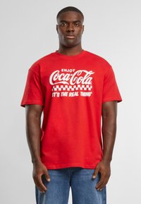 Rød bomulds t-shirt med "Coca-Cola" tekst i hvid og et ternet mønster. Korte ærmer, rund hals, afslappet pasform. Slidte denimjeans.
