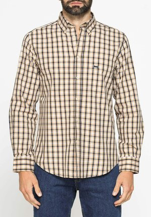 Uomo che indossa una camicia a maniche lunghe beige, bianca e nera a quadri con bottoni e jeans blu scuro, in piedi contro uno sfondo bianco.