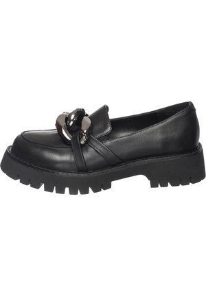 Sorte chunky platform loafers med oversized sølvkæde detaljer båret på bare fødder mod en lys baggrund.