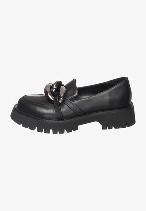 Mocassins à plateforme noire épaisse avec un détail de chaîne argentée surdimensionnée, portés pieds nus sur un fond clair.