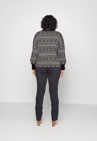 Personne aux cheveux bouclés portant un pull à motifs noir et blanc, un pantalon foncé et des chaussures à talons beiges, debout face à un mur blanc uni.