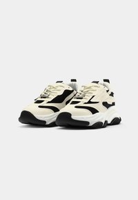 Zapatillas atléticas gruesas con parte superior de color crema, acentos de malla negra y una suela gruesa blanca y negra. Presenta cordones redondos y una lengüeta de tira.