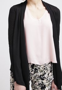 Schwarze offene Strickjacke über einer hellrosa V-Ausschnitt-Bluse, kombiniert mit floral gemusterten Hosen, die eine glatte Textur und einen fließenden Schnitt aufweisen.