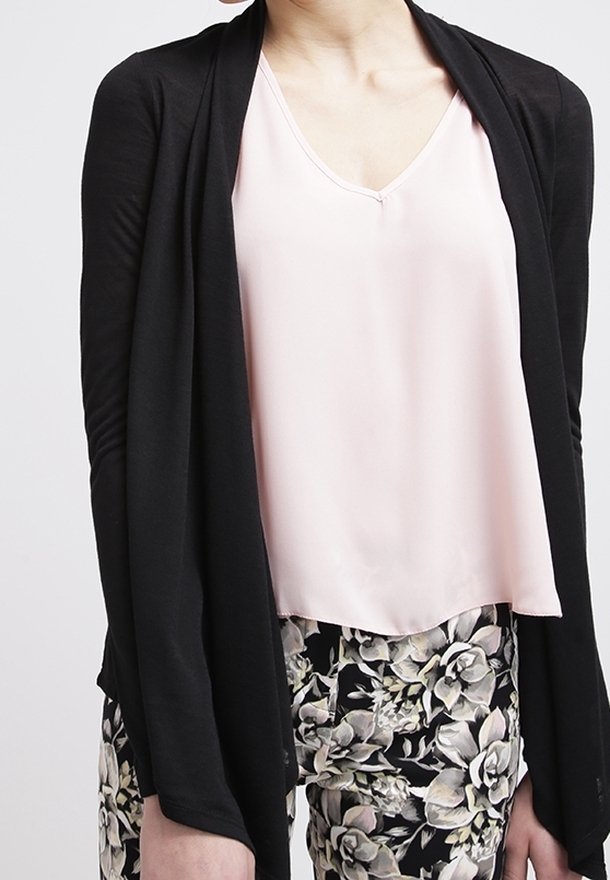 Schwarze offene Strickjacke über einer hellrosa V-Ausschnitt-Bluse, kombiniert mit floral gemusterten Hosen, die eine glatte Textur und einen fließenden Schnitt aufweisen.