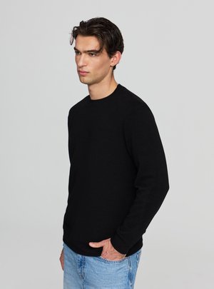 Felpa nera a maniche lunghe con tessuto strutturato e scollo rotondo, abbinata a jeans blu chiaro, mostrata in profilo laterale.