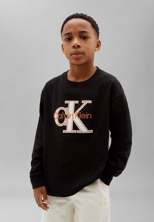 Fiú fekete Calvin Klein pulóverben és fehér nadrágban áll egy egyszerű szürke háttér előtt semleges arckifejezéssel.