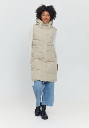 Beige Steppweste mit Kapuze, ausgestattet mit einem Frontreißverschluss, Seitentaschen und einer gesteppten Textur, kombiniert mit blauen weiten Jeans und schwarzen Stiefeln.