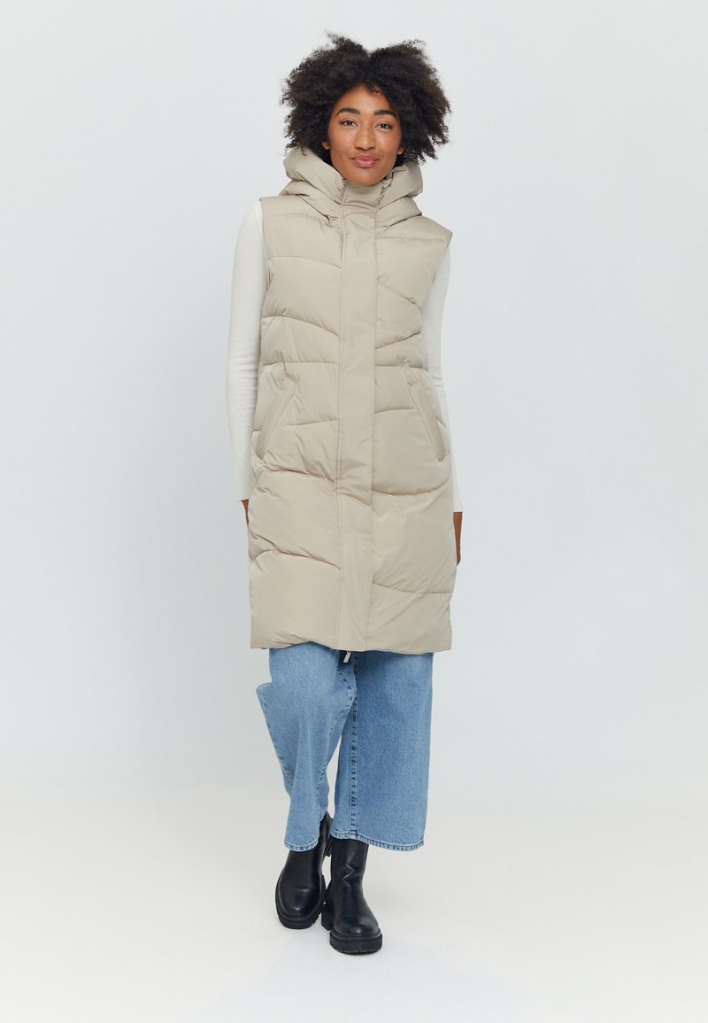 Beige puffer vest with a hood, featuring a front zipper, side pockets, and quilted texture, paired with blue wide-leg jeans and black boots.