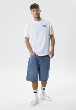 Mężczyzna stojący, ubrany w białą koszulkę z napisem "MEMENTO VIVERE", dżinsowe spodenki do kolan, białe trampki i białe skarpetki z czarnymi paskami.