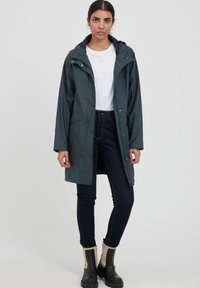 Waterdichte donkergroene jas met een capuchon, drukknoopsluiting en zijzakken, gecombineerd met een wit shirt, donkere spijkerbroek en zwarte laarzen.