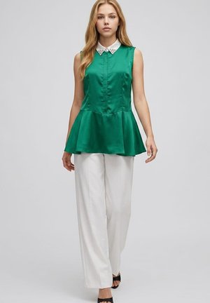 Mujer caminando hacia adelante con una blusa sin mangas verde con peplum, con cuello blanco y pantalones anchos blancos.