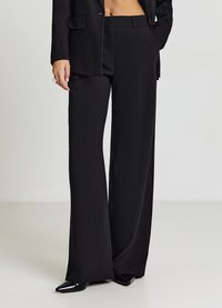 Schwarze, maßgeschneiderte Hose mit lockerem Sitz, vertikalem Nadelstreifenmuster und glattem Finish, kombiniert mit spitzen schwarzen Schuhen.