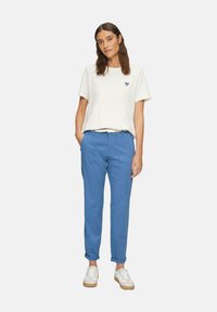 Witte katoenen t-shirt met een klein blauw hartontwerp, gecombineerd met blauwe chino's en witte sneakers met tan kleurige zolen, staande tegen een neutrale achtergrond.