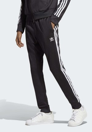 Pantalones deportivos negros con rayas blancas laterales, cintura elástica y bolsillos. Combinados con zapatillas blancas que cuentan con una puntera de goma.
