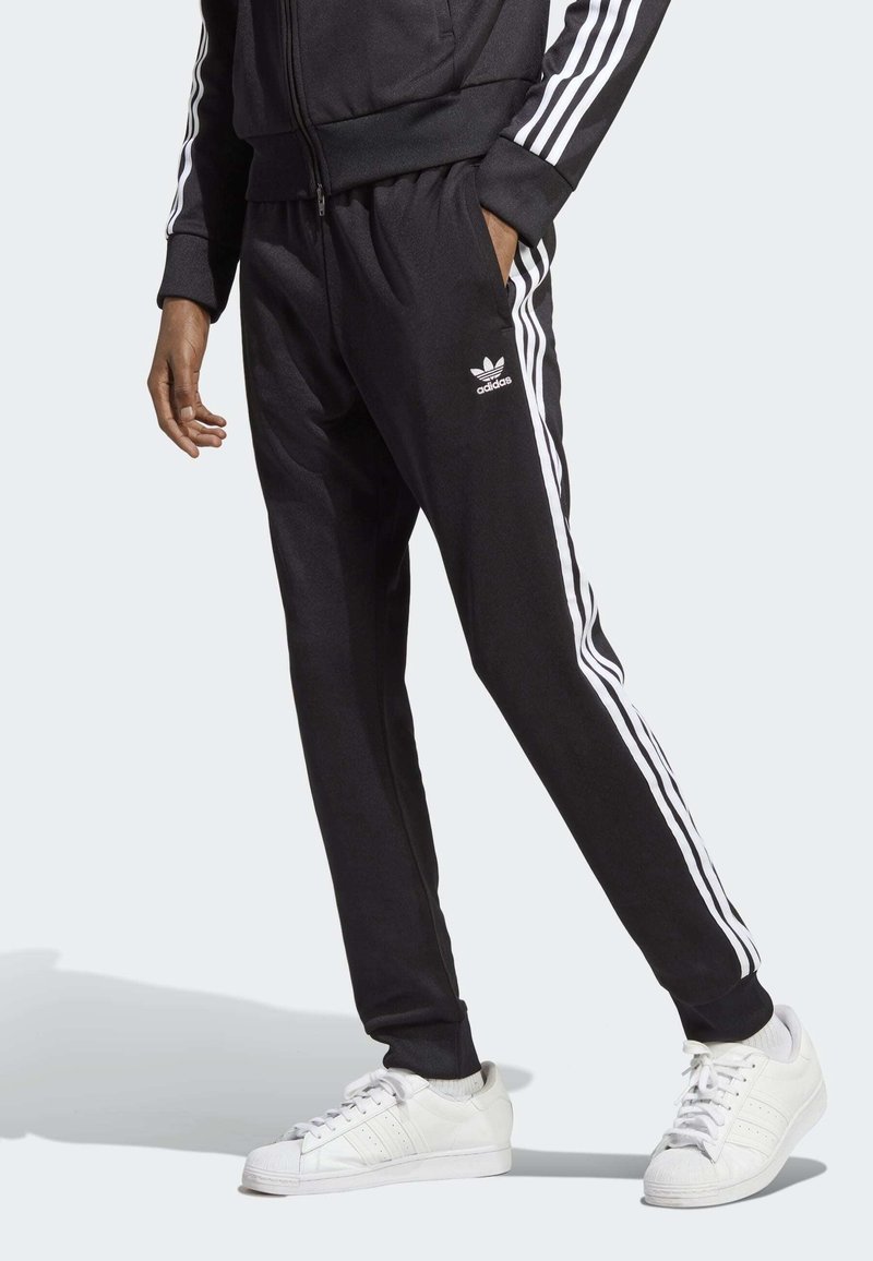 Pantalons de sport noirs avec des rayures blanches sur les côtés, taille élastique et poches. Associés à des baskets blanches avec une coque en caoutchouc.