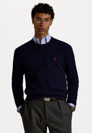 Marineblauwe kabelgebreide trui met ronde hals, voorzien van een klein rood logo. Draagt over een lichtblauw gestreept overhemd met kraag. Grijze broek en zichtbaar horloge.