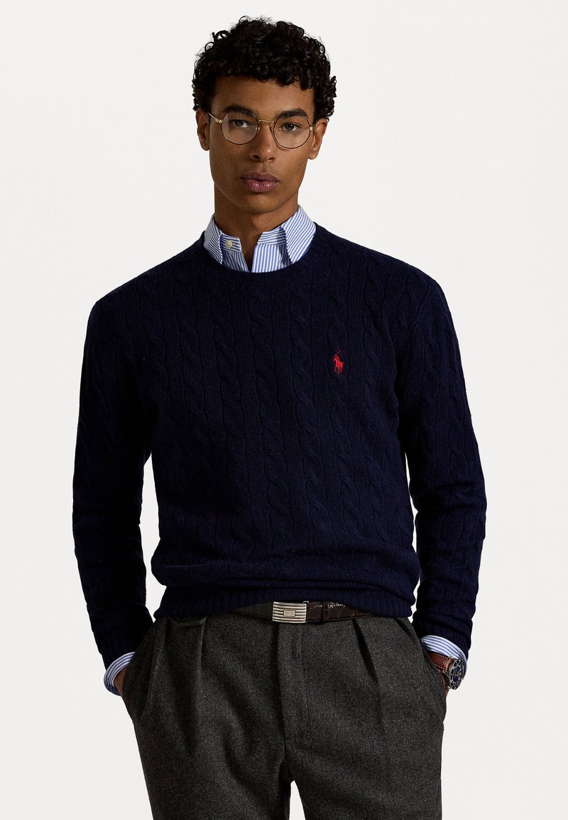 Navy Blauer Kabelstrickpullover mit rundem Halsausschnitt, versehen mit einem kleinen roten Logo. Getragen über einem hellblauen gestreiften Hemd mit Kragen. Graue Hose und eine Armbanduhr sind sichtbar.