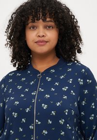 Veste zippée marine avec un motif floral composé de fleurs bleues et de feuilles vertes, fabriquée à partir d'un tissu lisse au toucher doux.