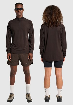 Bruine lange mouwen, met rits, van textuurstof, gecombineerd met bruine shorts met een logo. Model draagt donkere zonnebrillen en dikke schoenen.