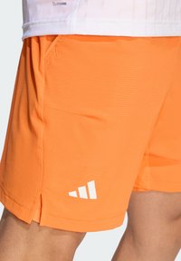 Φωτεινά πορτοκαλί αθλητικά σορτς από υφασμάτινη υφή, με λευκό λογότυπο και μικρές πλαϊνές σχισμές για ευκολία στην κίνηση.