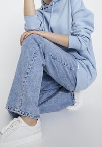 Personne assise jambes croisées portant un sweat à capuche bleu clair, un jean bleu délavé et des baskets blanches sur un fond blanc uni.