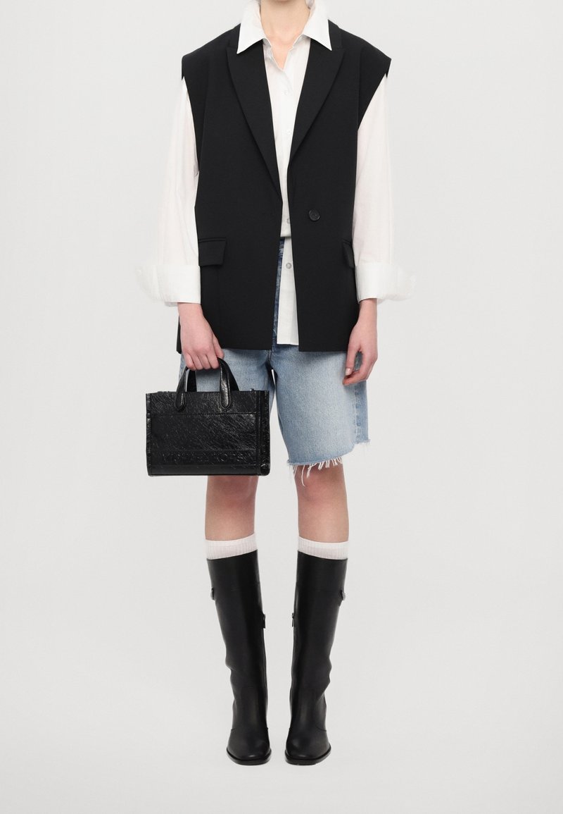 Personne portant un blazer noir sans manches, une chemise blanche, un short en jean à hauteur des genoux, des bottes noires hautes jusqu'aux genoux, et tenant un sac à main noir.