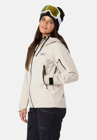 Giacca softshell beige chiaro con dettagli neri, tasche con zip e cappuccio regolabile, abbinata a pantaloni da neve neri e un berretto in maglia.