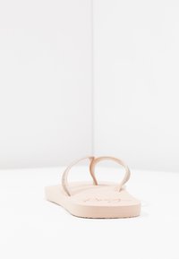 Flip-flop bej cu o talpă texturată, având două curele subțiri de culoare aurie roz, împodobite cu mici detalii strălucitoare. Logo-ul este vizibil pe gamă.