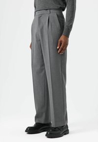 Pantalon plissé gris en tissu mélangé laine. Caractérisé par une coupe ajustée, une taille haute et des poches. Porté avec des chaussures noires épaisses.
