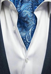 Sininen paisley-silkki solmio valkoisen paitakauluksen alla; paitapainikkeet näkyvissä; tumma, teksturoidun kangaspuvun reuna.
