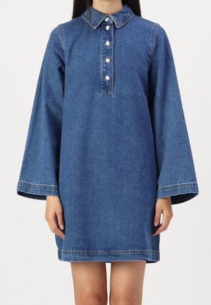 Kobieta ubrana w długą sukienkę z niebieskiego denimu z szerokimi rękawami i przednim panelem zapinanym na cztery guziki, stojąca na tle białego tła.