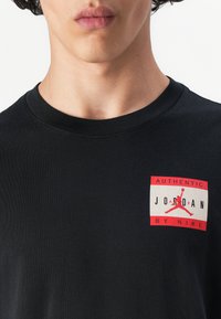 Sort bomulds t-shirt med rund hals. Har et rødt og hvidt firkantet logo på venstre brystområde, der står "AUTHENTIC JORDAN BY NIKE."