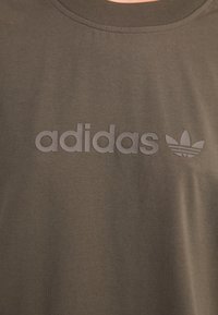 Tamsiai žalias medvilnės marškinėliai su iškilniu pilku adidas logotipu ir trimis juostelėmis. Suapvalintas iškirpė ir minkšta tekstūra.