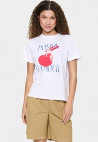Balta medvilninė marškinėliai su dideliu raudono obuolio grafiku ir tekstu "POMME D'AMOUR" mėlyna spalva. Trumpomis rankovėmis, laisvas kirpimas ir apvaliu kaklu.