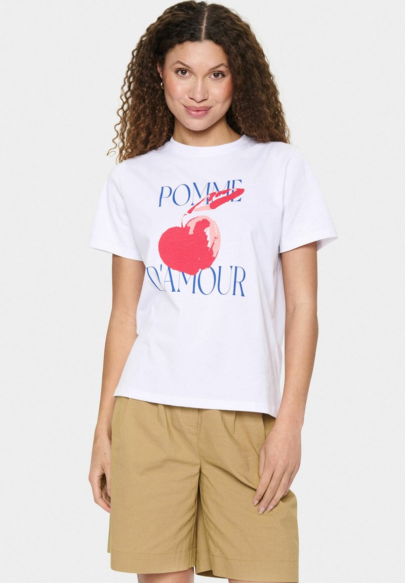 Balta medvilninė marškinėliai su dideliu raudono obuolio grafiku ir tekstu "POMME D'AMOUR" mėlyna spalva. Trumpomis rankovėmis, laisvas kirpimas ir apvaliu kaklu.