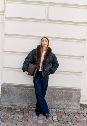 Femme debout contre un mur blanc, vêtue d’une veste noire à col en fourrure, d’un jean large foncé, de chaussures noires pointues et tenant une pochette marron.