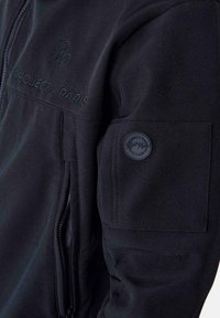 Giacca nera con una texture liscia, dotata di una tasca con zip e di un patch rotondo con il logo sulla manica. Testo ricamato sopra la tasca.