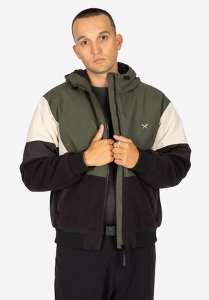 Mann mit kurzen Haaren, der eine grüne, schwarze und beige Jacke trägt und mit beiden Händen den Reißverschlussbereich vor einem weißen Hintergrund hält.