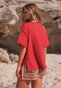 T-shirt rouge oversize avec manches retroussées, associée à un short multicolore à motifs agrémenté d'accents en crochet, sur fond rocheux.